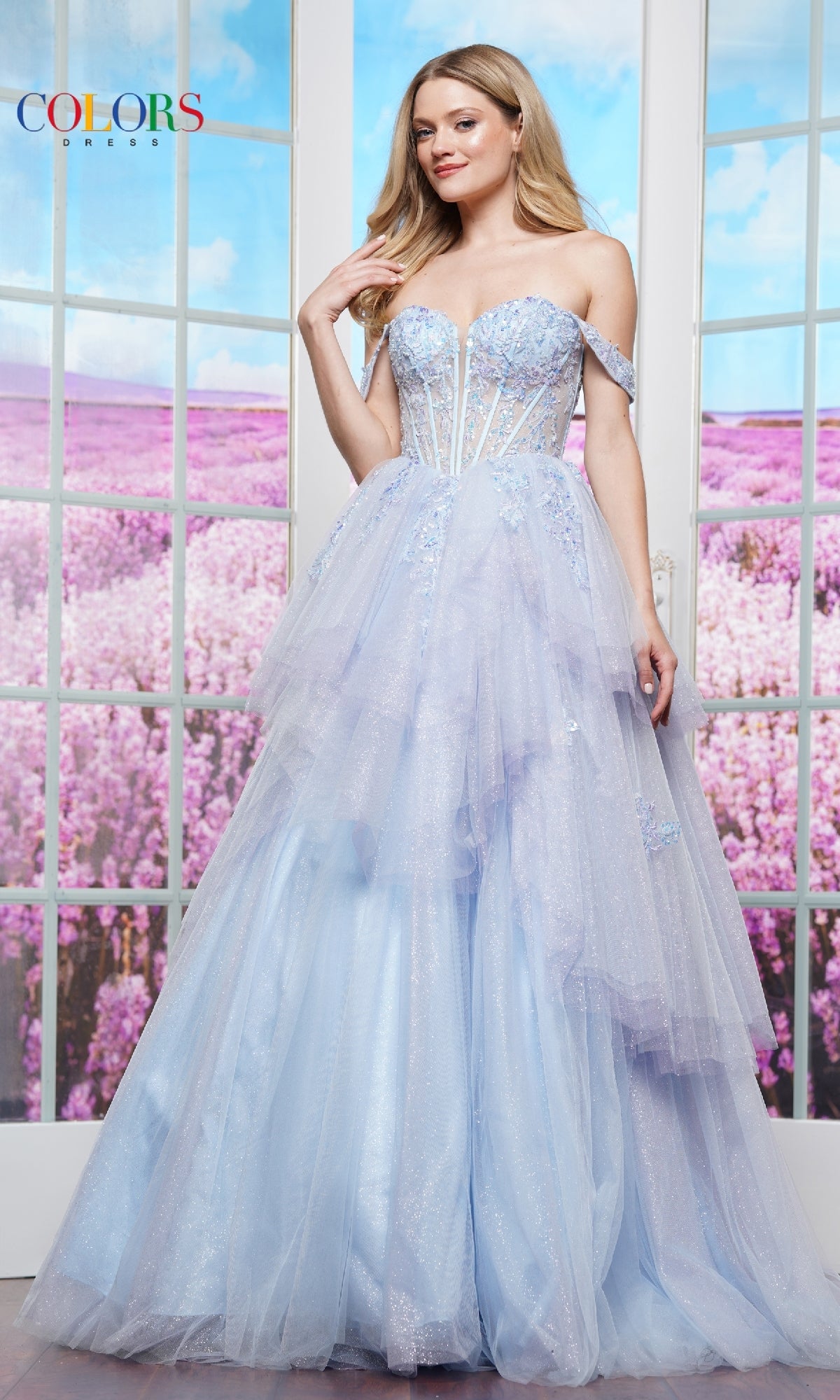 A woman wearing the Off-Shoulder Long Prom Ball Gown: Colors Dress 3534 poses in front of glass doors with a lavender field in the background.